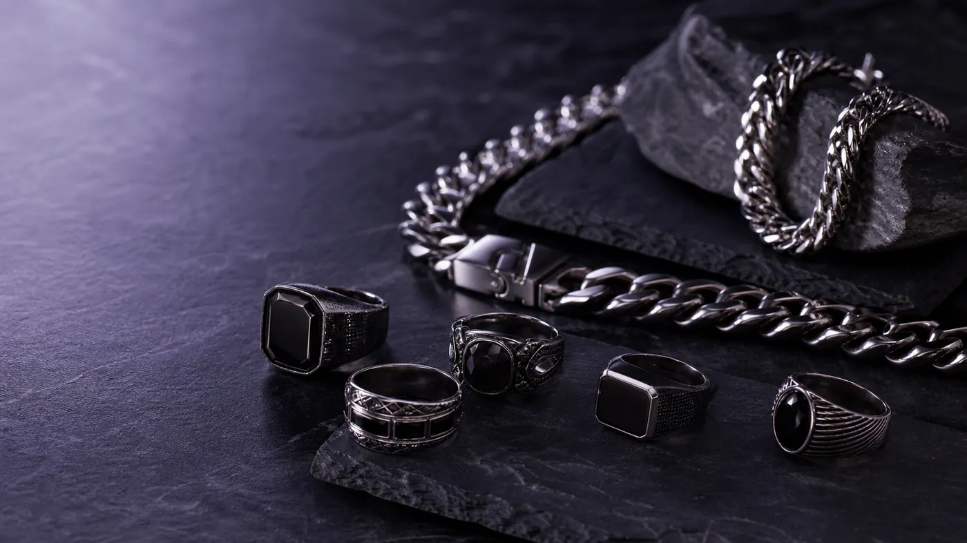 Gunmetal signet rings and heavy silver cuban-link chains on dark slate under violet light